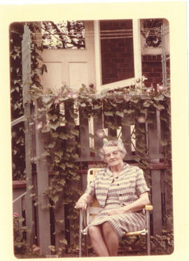 Photograph of an older woman sitting in a lawn chair in front of a house and a vine-covered fence