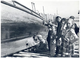 Photograph of a man looking at a hole in the keel of a salvaged boat, sitting on a beach at Seal ...