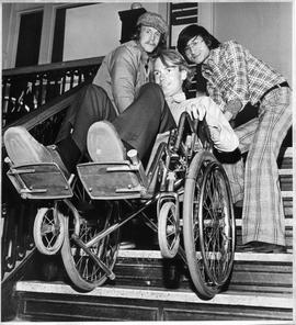 Photograph of Duncan Coates and Alex Fok lifting Larry Richards in a wheelchair