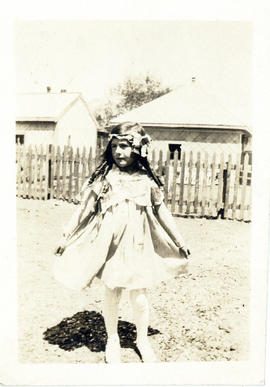 Photograph of a young girl standing in a fenced yard, holding her skirt and wearing ribbons in he...