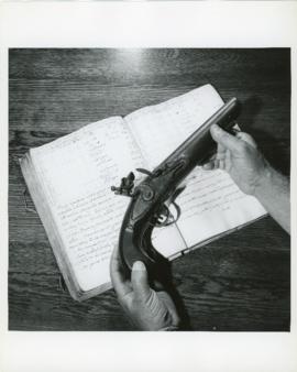 Photograph of a flintlock pistol held by Thomas Head Raddall over the logbook of the Spanish ship...
