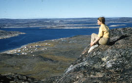 Photograph of Barbara Hinds sitting on a mountain overlooking Cape Dorset, Northwest Territories