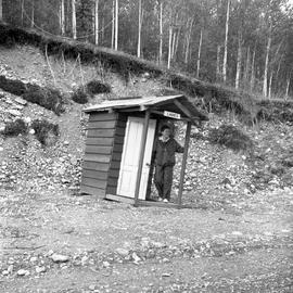 Photograph of Barbara Hinds standing in front of an outhouse in the Yukon