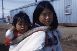 Photograph of an unidentified woman in a white coat carrying a baby on her back