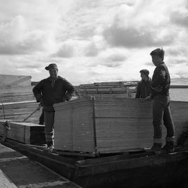 Photograph of three men with supplies on a barge on the Koksoak River