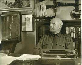 Photograph of Thomas Head Raddall sitting at his desk in his study with a bookshelf and decorativ...