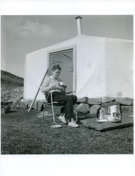 Photograph of Barbara Hinds sitting outside a tent and drinking tea