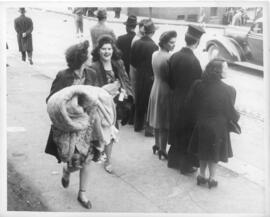 Photograph of two women carrying looted goods, while others watch events on the street during the...