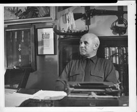 Photographic negative of Thomas Head Raddall sitting at the desk in his study with a bookshelf, d...