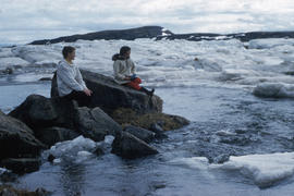 Photograph of Barbara Hinds and another woman sitting on rocks by the water in Frobisher Bay, Nor...