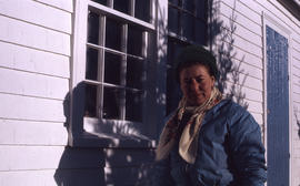 Photograph of Barbara Hinds standing by the window of a white house