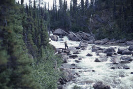 Photograph of an unidentified person standing by river rapids near Voisey's Bay, Newfoundland and...