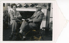 Photograph of Lloyd MacInnis interviewing Thomas Head Raddall, who is holding a book and wearing ...
