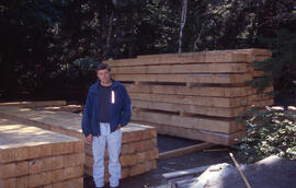 Photograph of an unidentified person standing in front of a stack of squared timber from trees da...