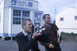 Photograph of a man with a video camera and another man with a cigar in front of a shop called "E...