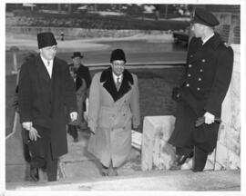 Photograph of a naval officer and two men in civilian dress walking up a set of outside stairs