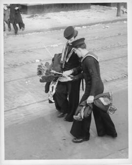 Photograph of two sailors examining looted goods, including a feather duster and fur vest, on VE-...