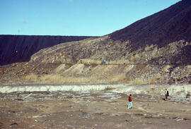 Photograph of unidentified researchers standing at the base of slag heaps at the Coniston site, n...