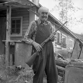 Photograph of Pete Brady standing in front of his house with a rifle