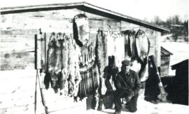 Photograph of Gordon Longevin posing outside in the winter next to a wall of mounted furs in Bisc...