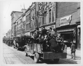 Photograph of servicemen riding in the back of a truck in a convoy of marching military personnel...