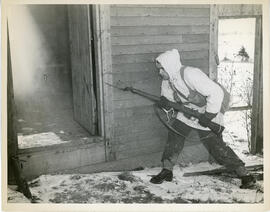 Photograph of an unidentified soldier carrying a bayonet outside a small cabin during winter infa...