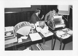 Photograph of two unidentified people with a table full of audiovisual equipment
