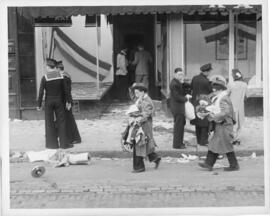 Photograph of civilian looters carrying stacks of clothing past the smashed shop windows during t...