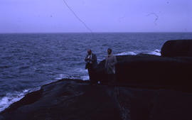 Photograph of two unidentified men standing on giant rocks by the water